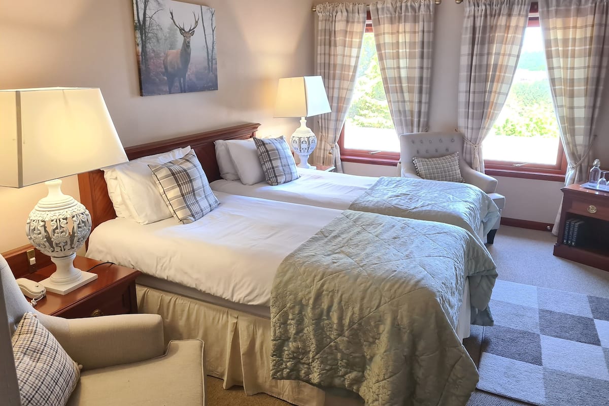 A hotel room with two beds, a chair, a lamp, a painting, and a window with curtains.
