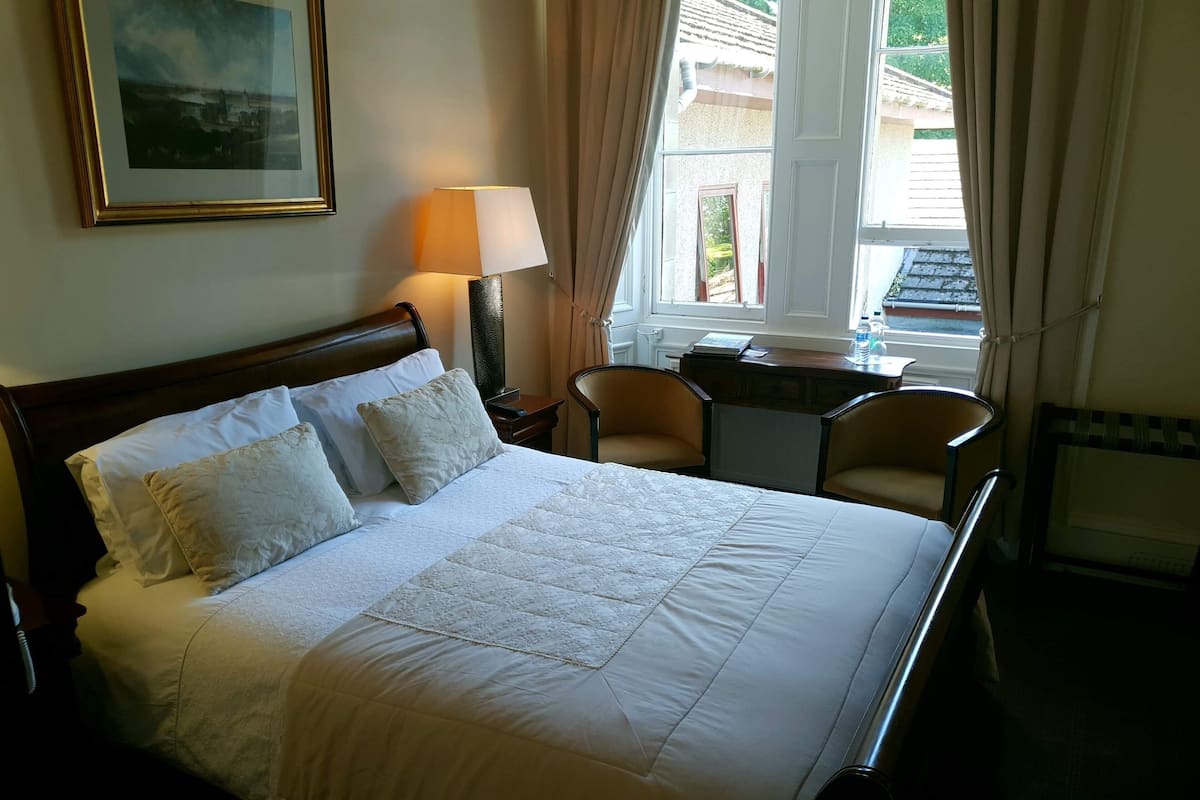 A neatly made bed with a quilt, a bedside lamp, and a chair near a window with curtains.