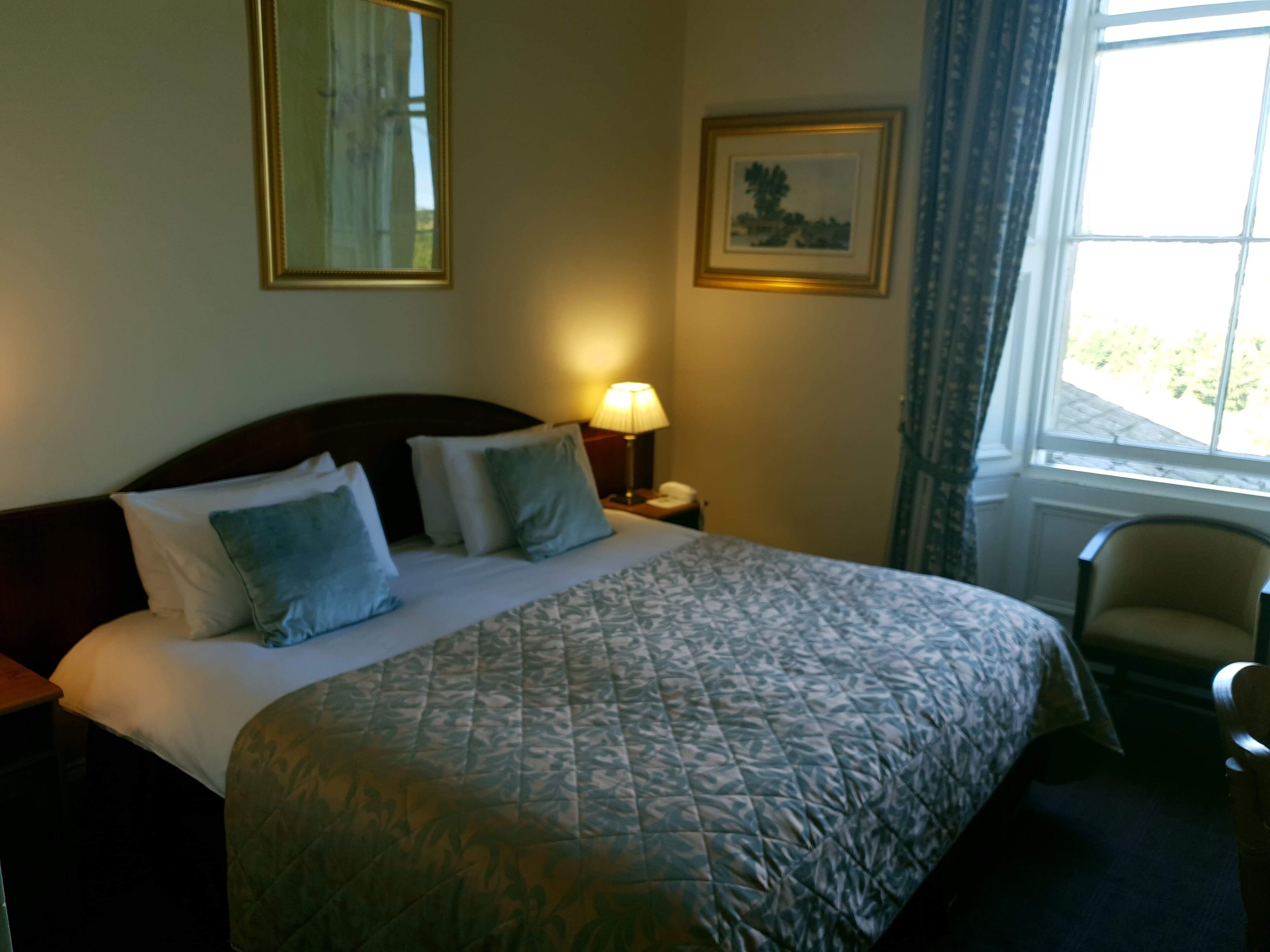Una habitación de hotel con una cama grande, una mesita de noche con lámpara, una silla y una ventana con cortinas.