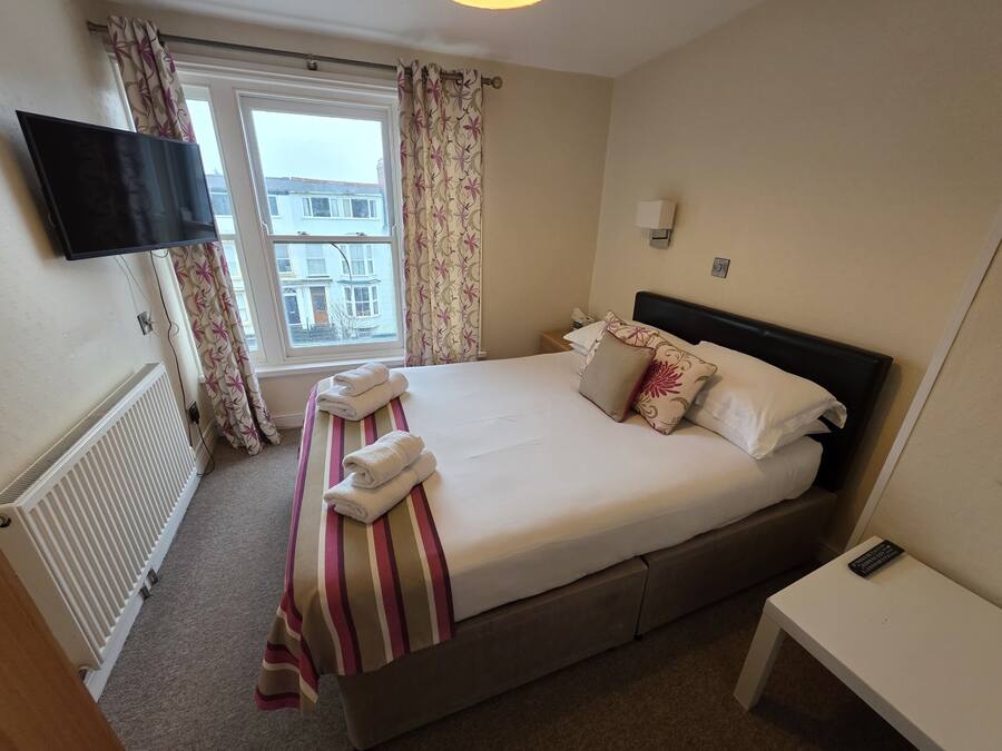 A hotel room with a bed, a TV mounted on the wall, a window with curtains, a bedside table, and a radiator.