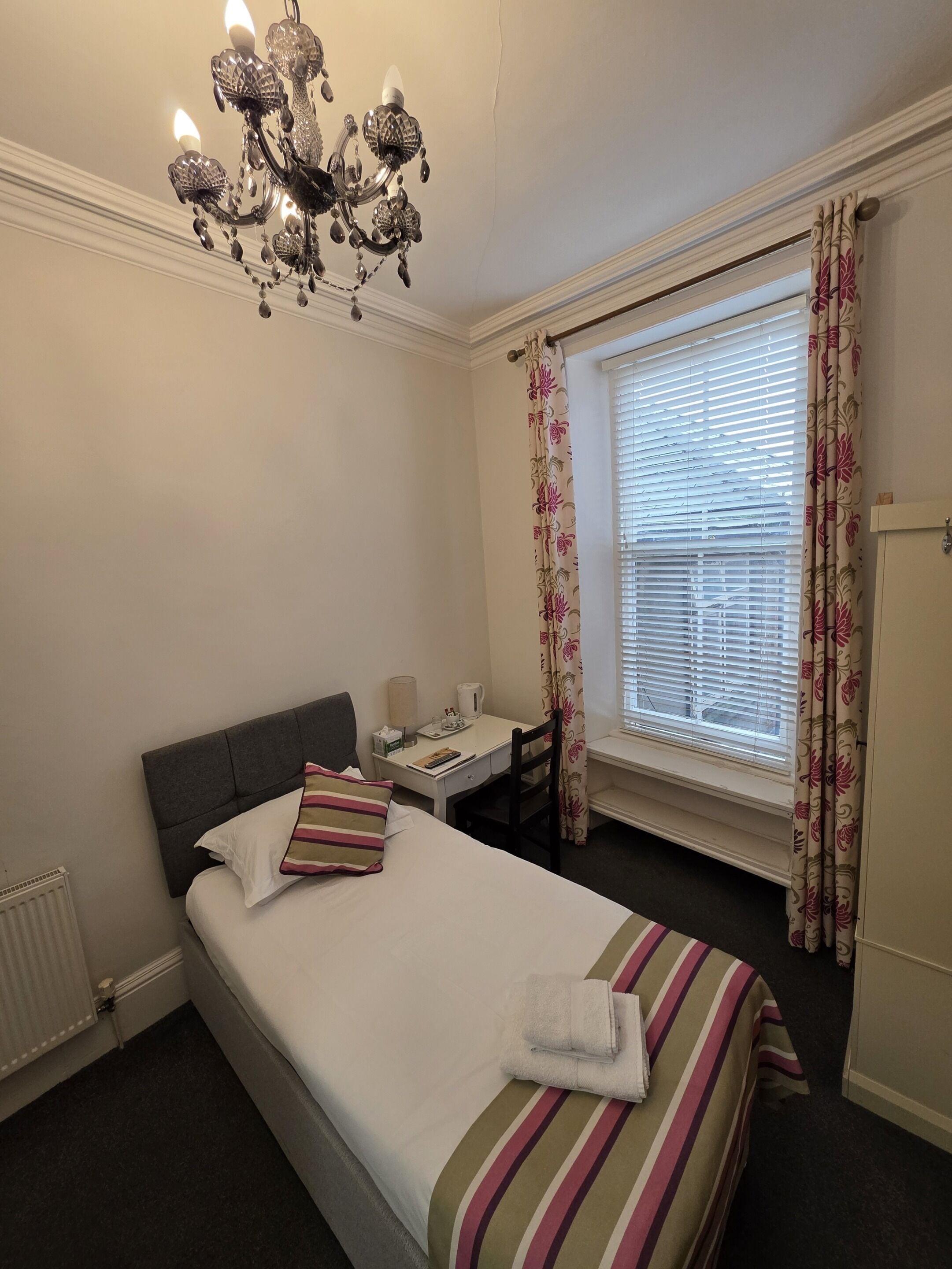 A hotel room with a bed, a desk, a chair, a radiator, a window with curtains, and a chandelier.