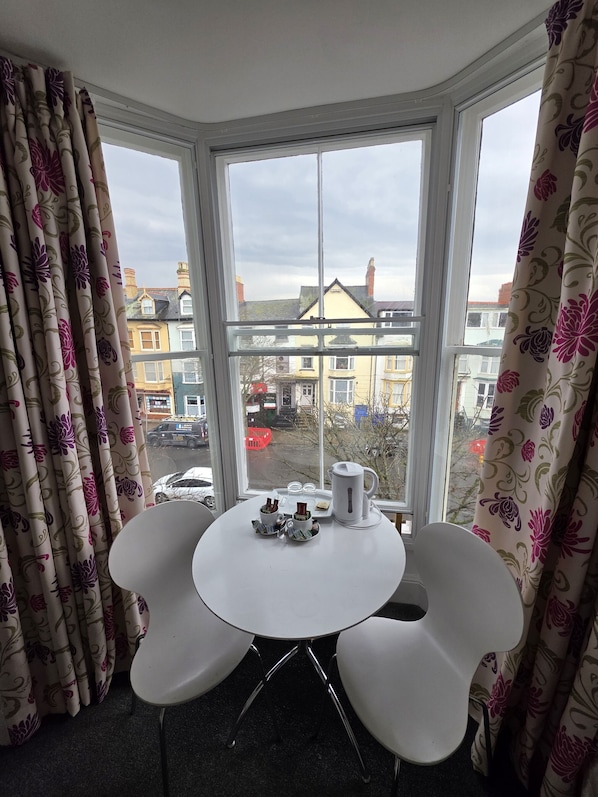 Una habitación con ventanal, dos sillas blancas, una mesita con bandeja y cortinas con estampado floral.