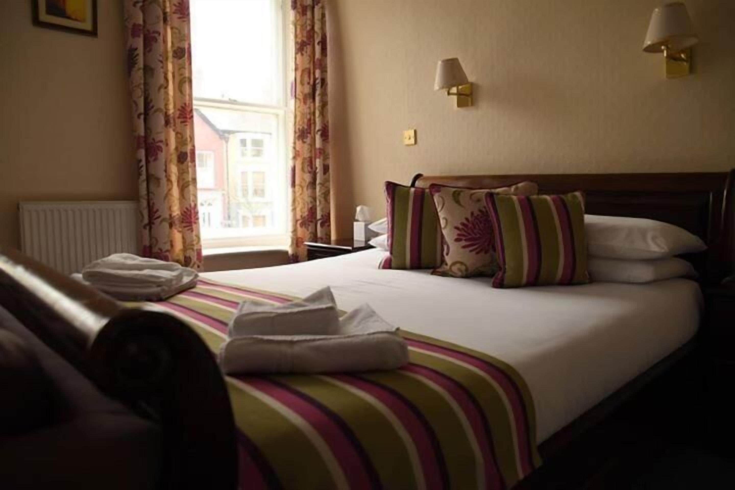 Habitación de hotel con cama, almohadas, mesita de noche, lámpara y ventana con cortinas.