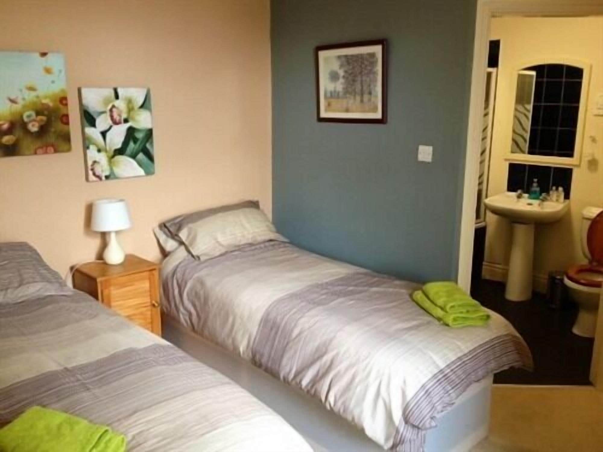 Une chambre avec deux lits, une table de chevet avec une lampe, un tableau encadré au mur et une salle de bain visible à travers une porte ouverte.