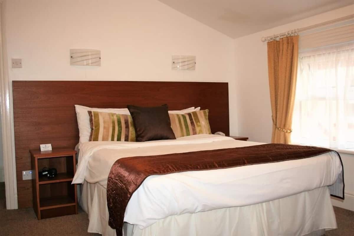 Een slaapkamer met een houten hoofdbord, een nachtkastje met een lamp, een raam met gordijnen en een bed met een wit-bruine dekbedsprei.