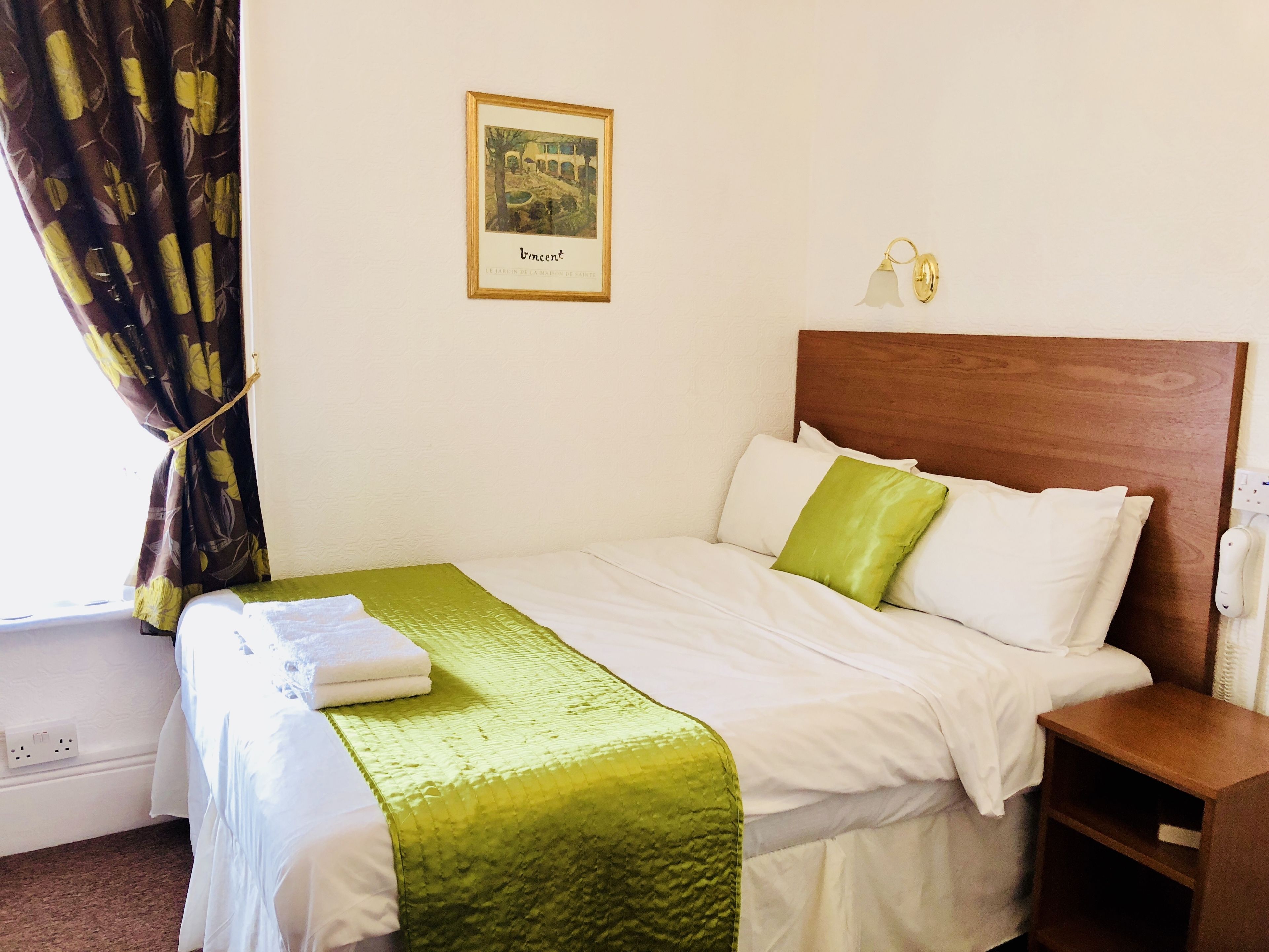 A hotel room with a neatly made bed, a green throw blanket, and a framed picture on the wall.