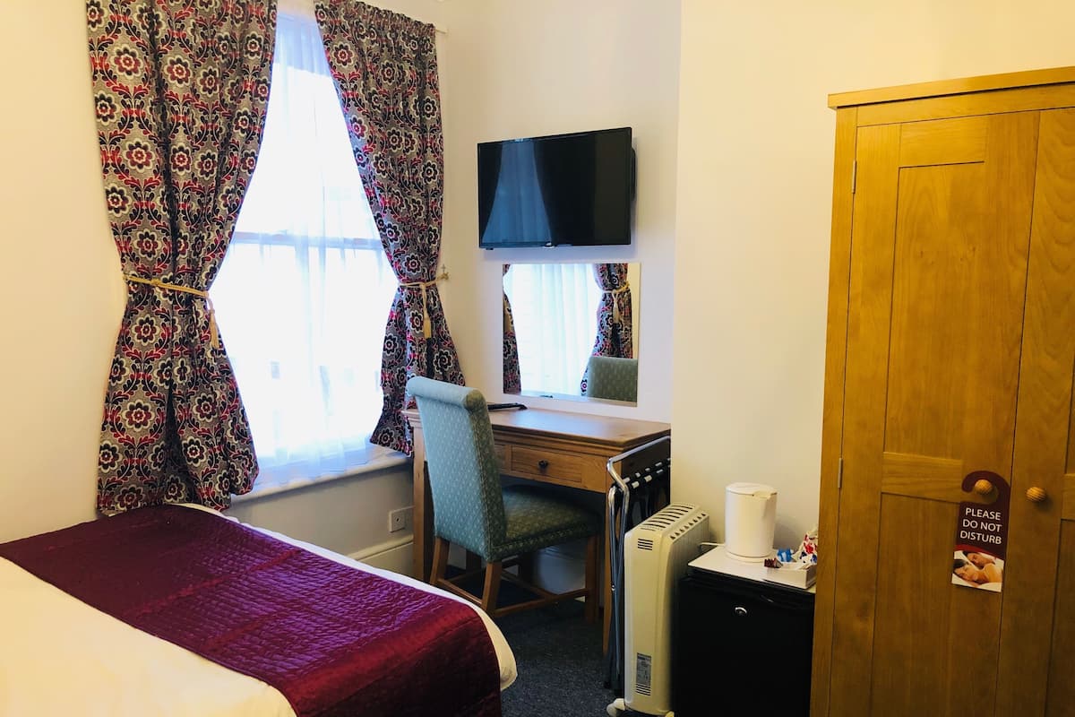 Een hotelkamer met een bed, een bureau met stoel, een televisie, een radiator en een deur met een bordje.