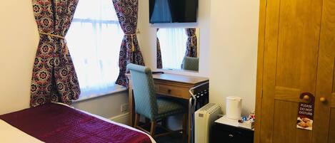 Habitación de hotel con una cama, un escritorio con silla, un televisor, un radiador y una puerta con cartel.
