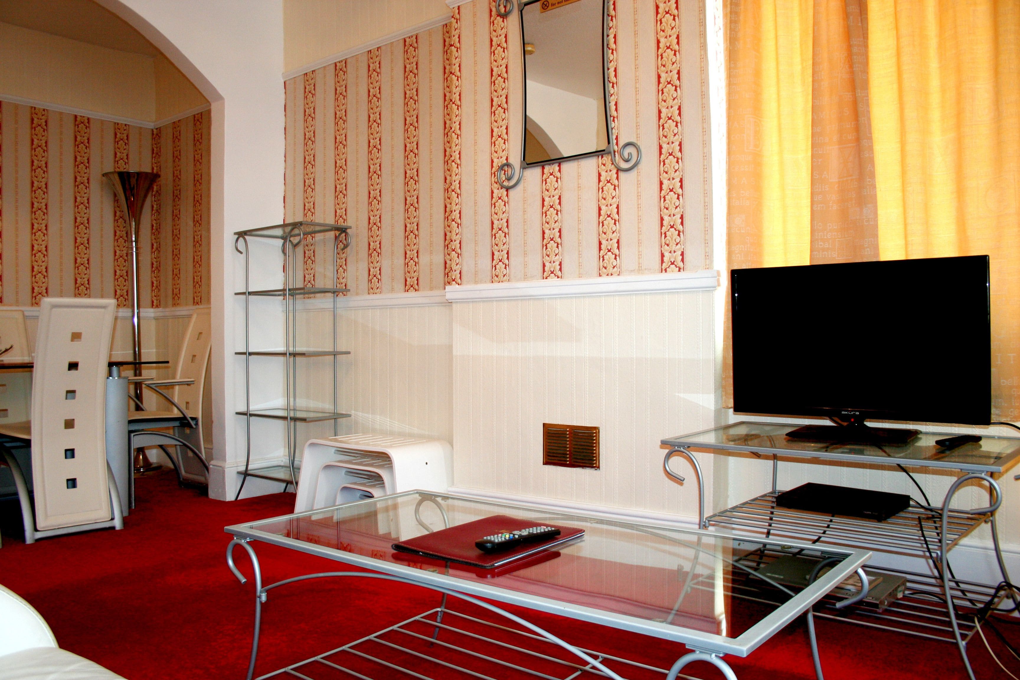 Een hotelkamer met een glazen tafel, een rode map, een zwarte afstandsbediening, een televisie en een spiegel aan de muur.