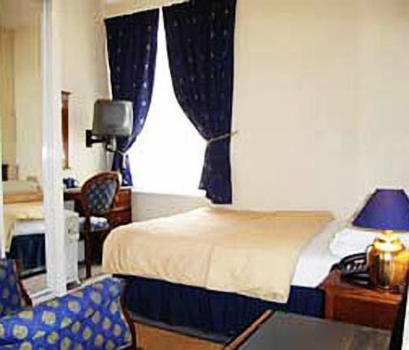 Quarto de hotel com cama, poltrona, escrivaninha, luminária e televisão.