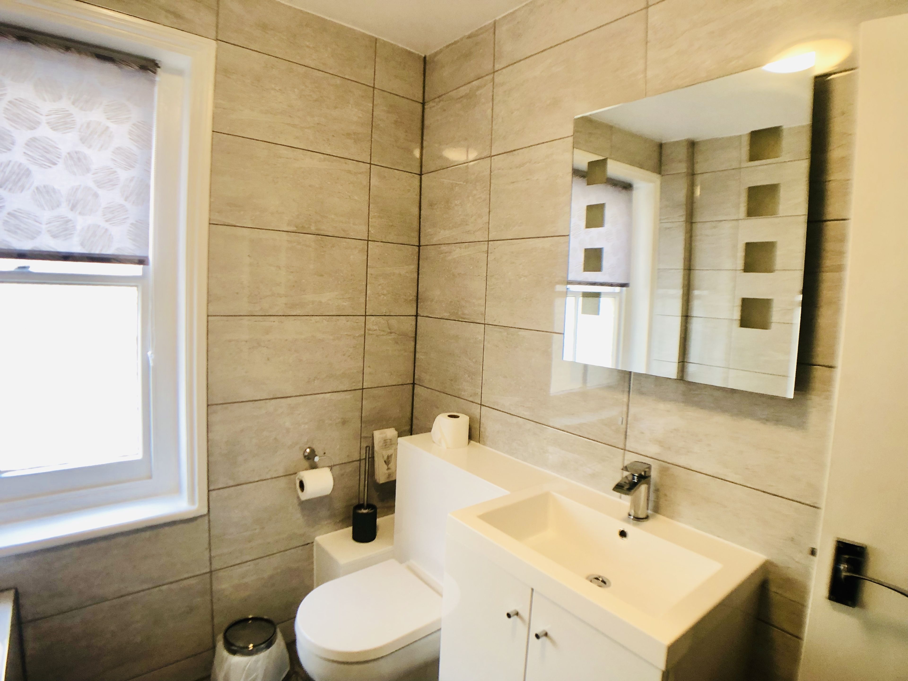 A modern bathroom with beige tiles, a white sink, a toilet, and a mirror.