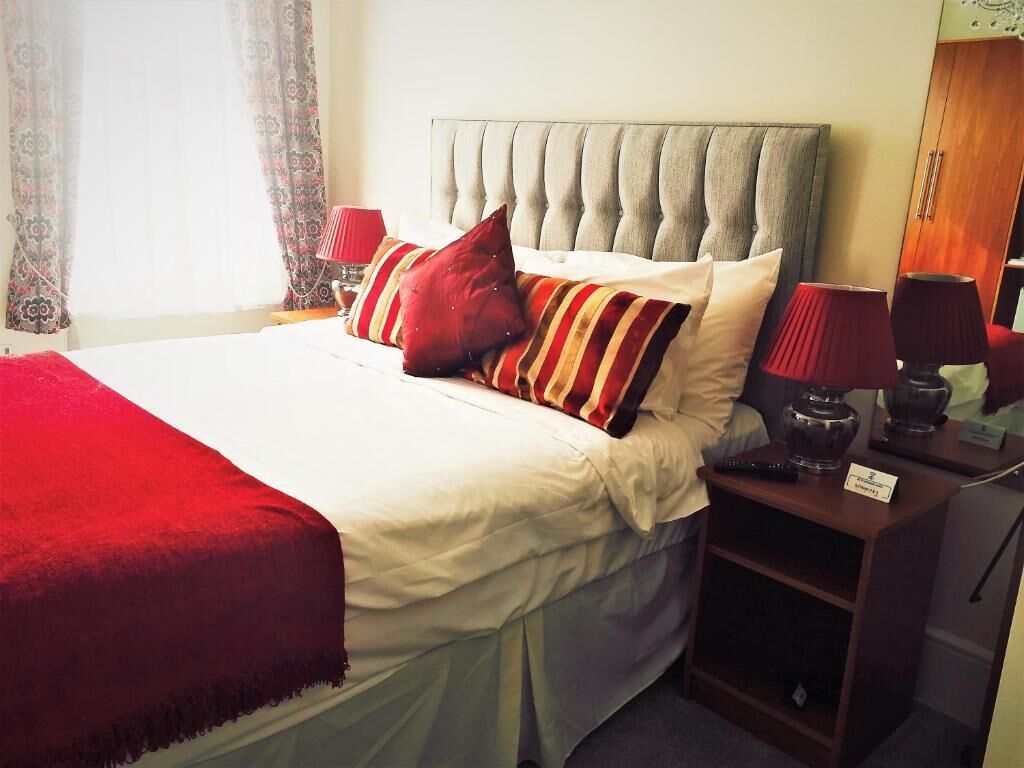 Een net opgemaakt bed met een rode deken, een grijs gestoffeerd hoofdbord en meerdere kussens. Er staat een nachtkastje met een lamp en een klein plankje.
