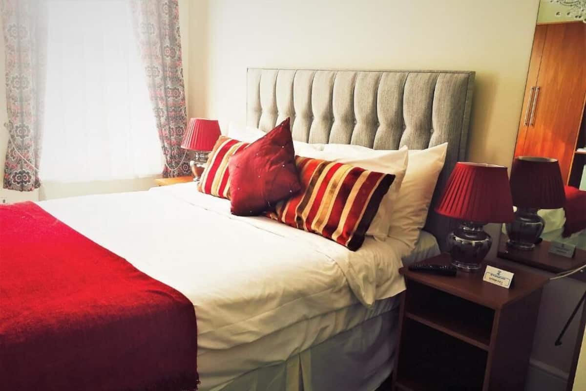 Een net opgemaakt bed met een rode deken, een grijs gestoffeerd hoofdbord en meerdere kussens. Er staat een nachtkastje met een lamp en een klein plankje.