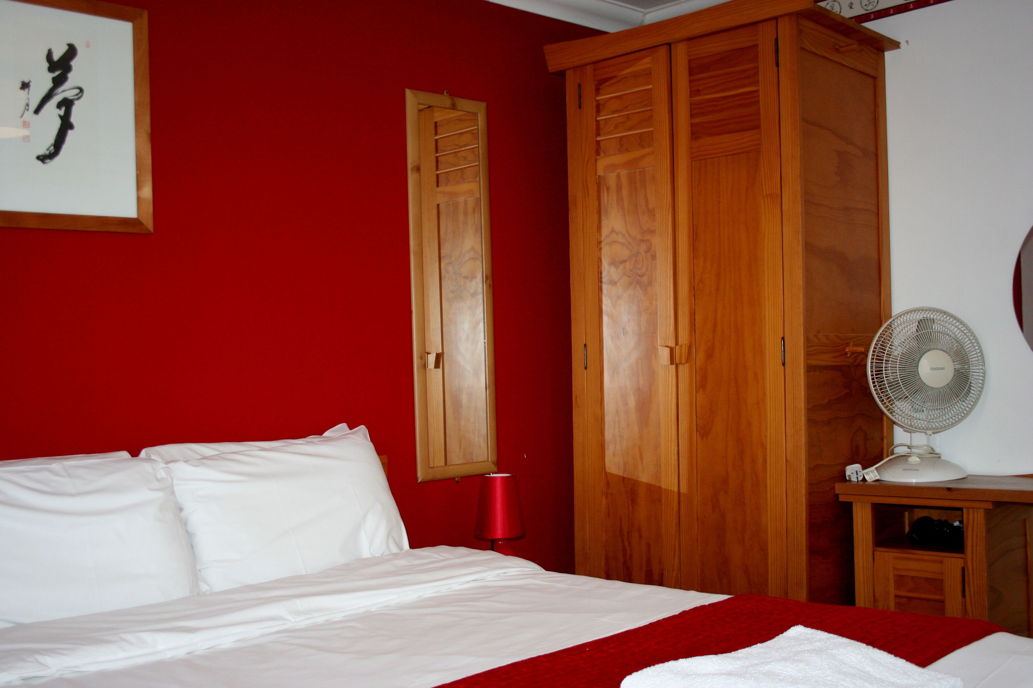 A hotel room with a bed, wooden wardrobe, a red wall, and a white fan.