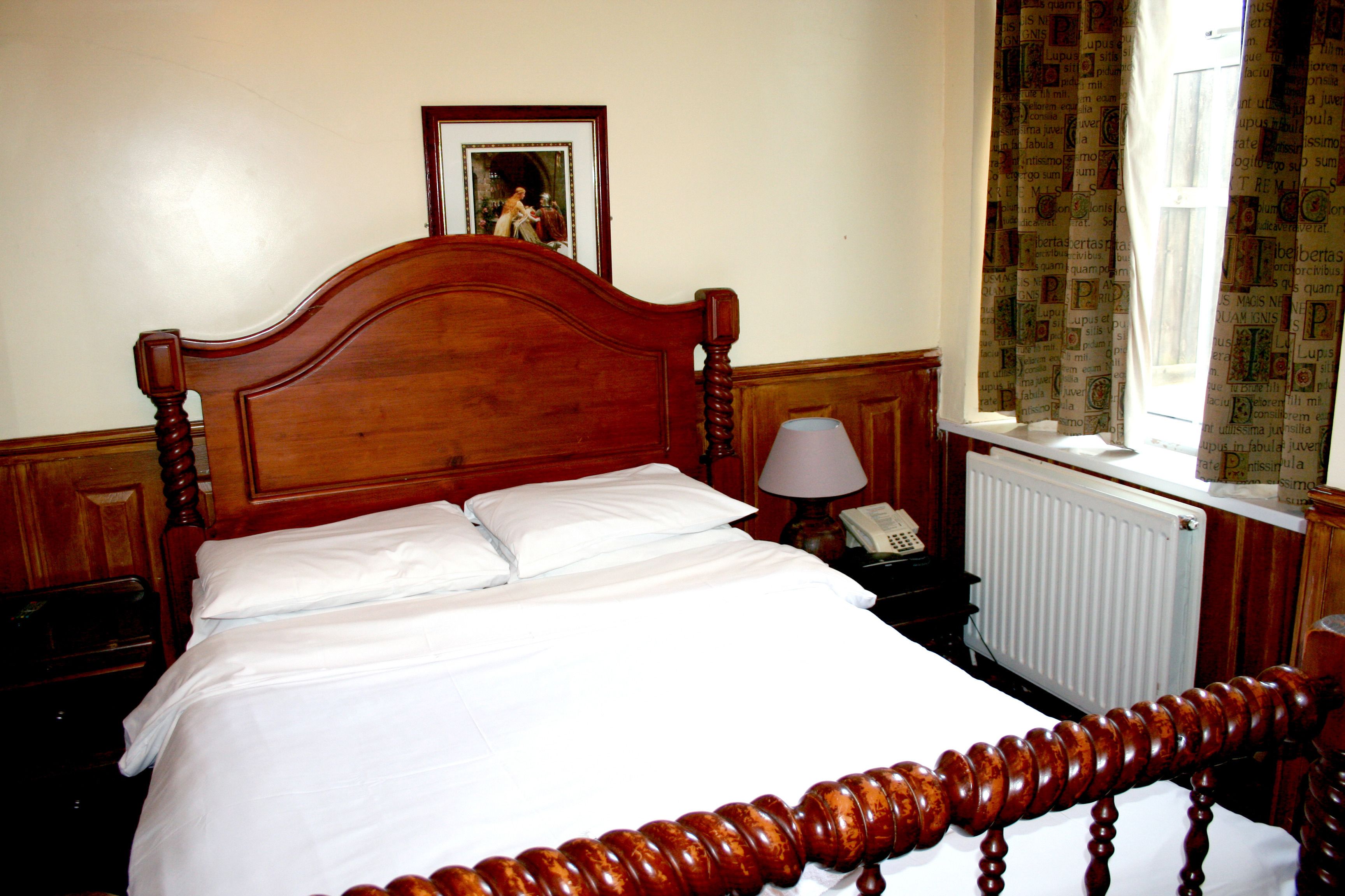 Una cama con dosel de madera con ropa de cama blanca, una lámpara de noche, un cuadro enmarcado en la pared y una ventana con cortinas estampadas.