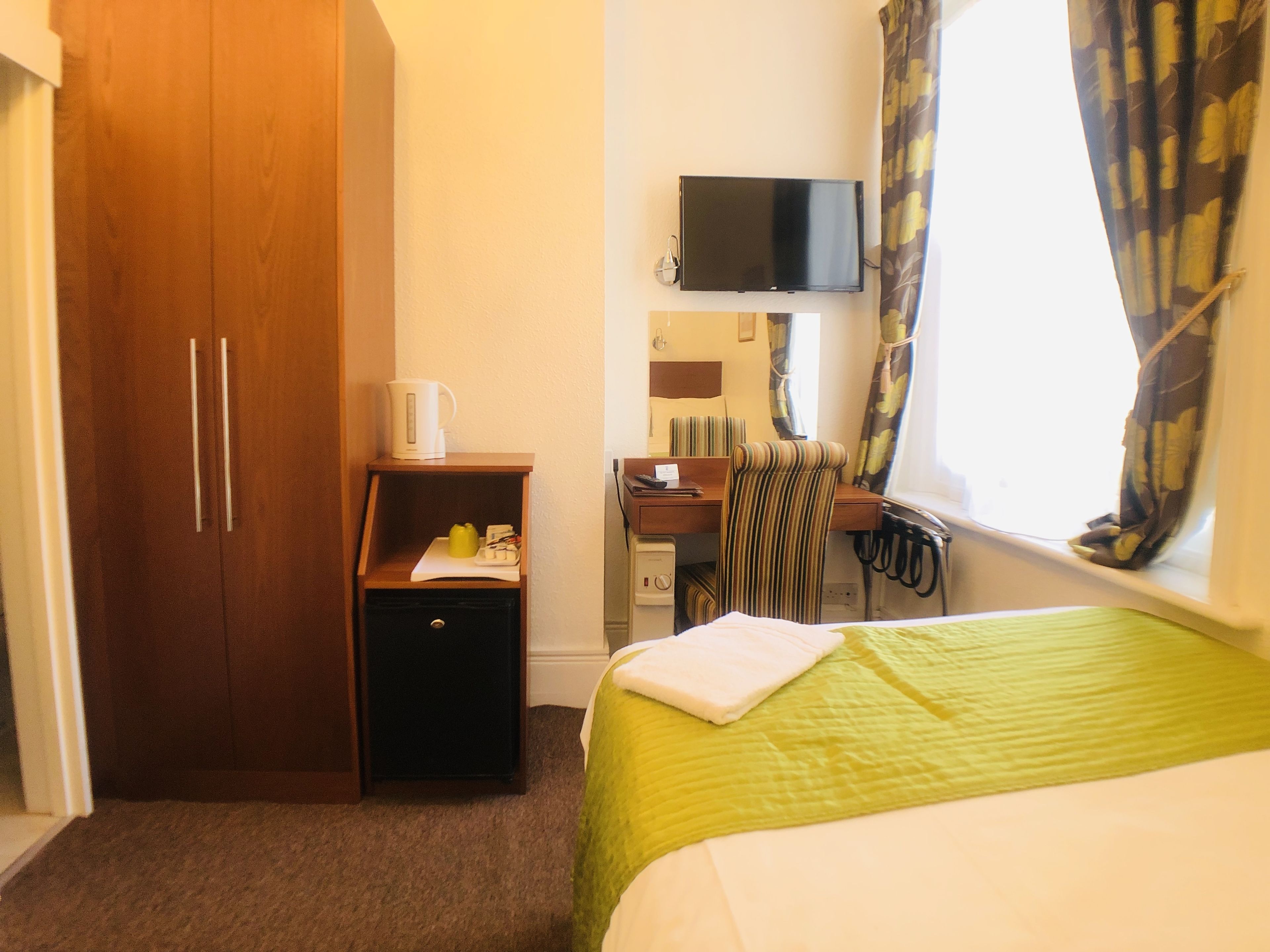 Ein Hotelzimmer mit Bett, Schreibtisch mit Stuhl, Fernseher, Kleiderschrank und einem Fenster mit Vorhängen.