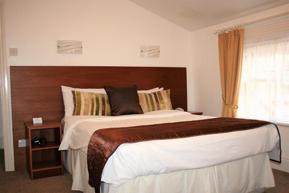 Une chambre à coucher avec une tête de lit en bois, une table de chevet avec une lampe, une fenêtre avec des rideaux et un lit recouvert d’une couvre-lit blanche et marron.