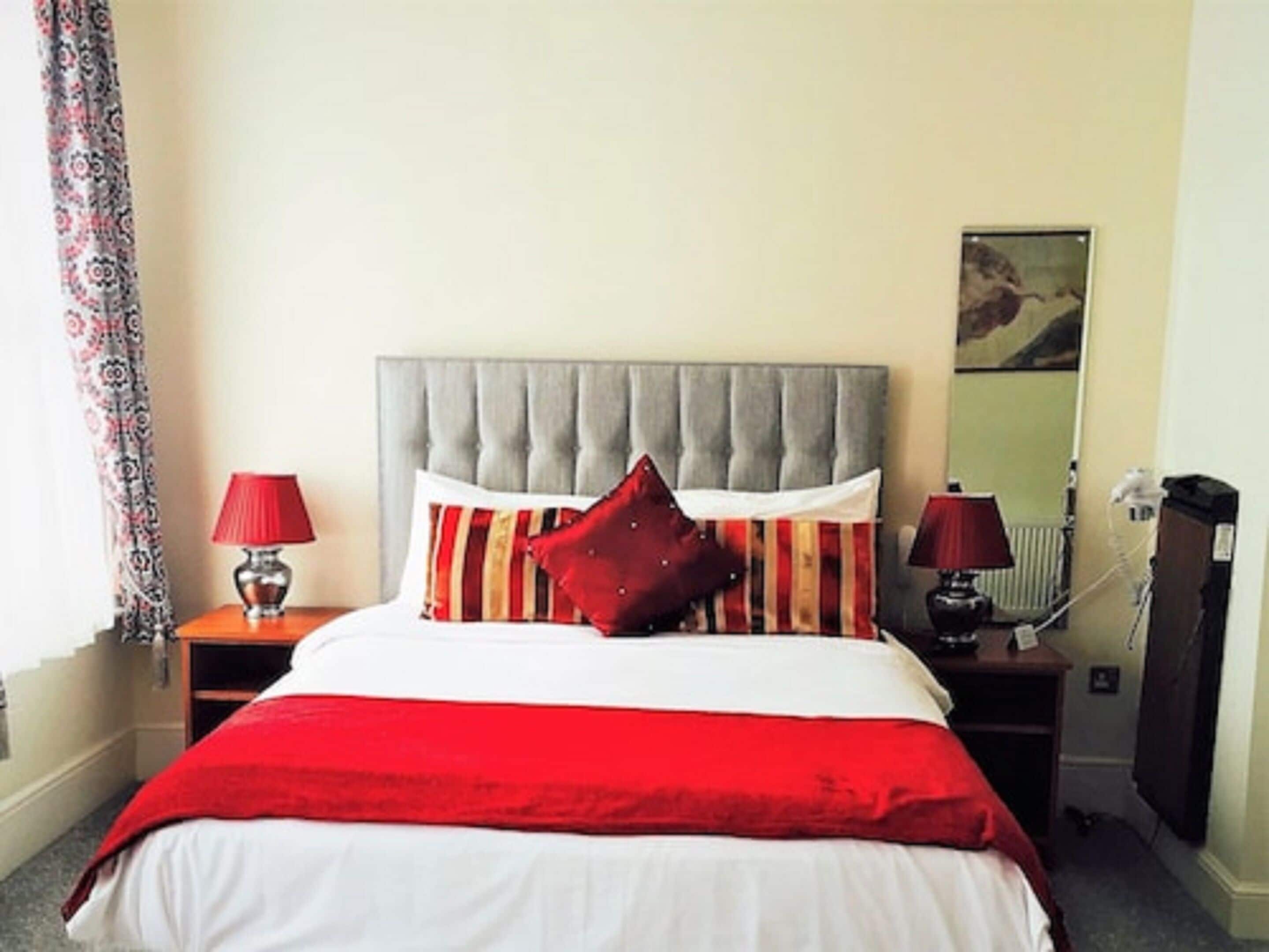 Habitación de hotel con una cama grande, dos mesitas de noche con lámparas, una manta roja y una cortina estampada.