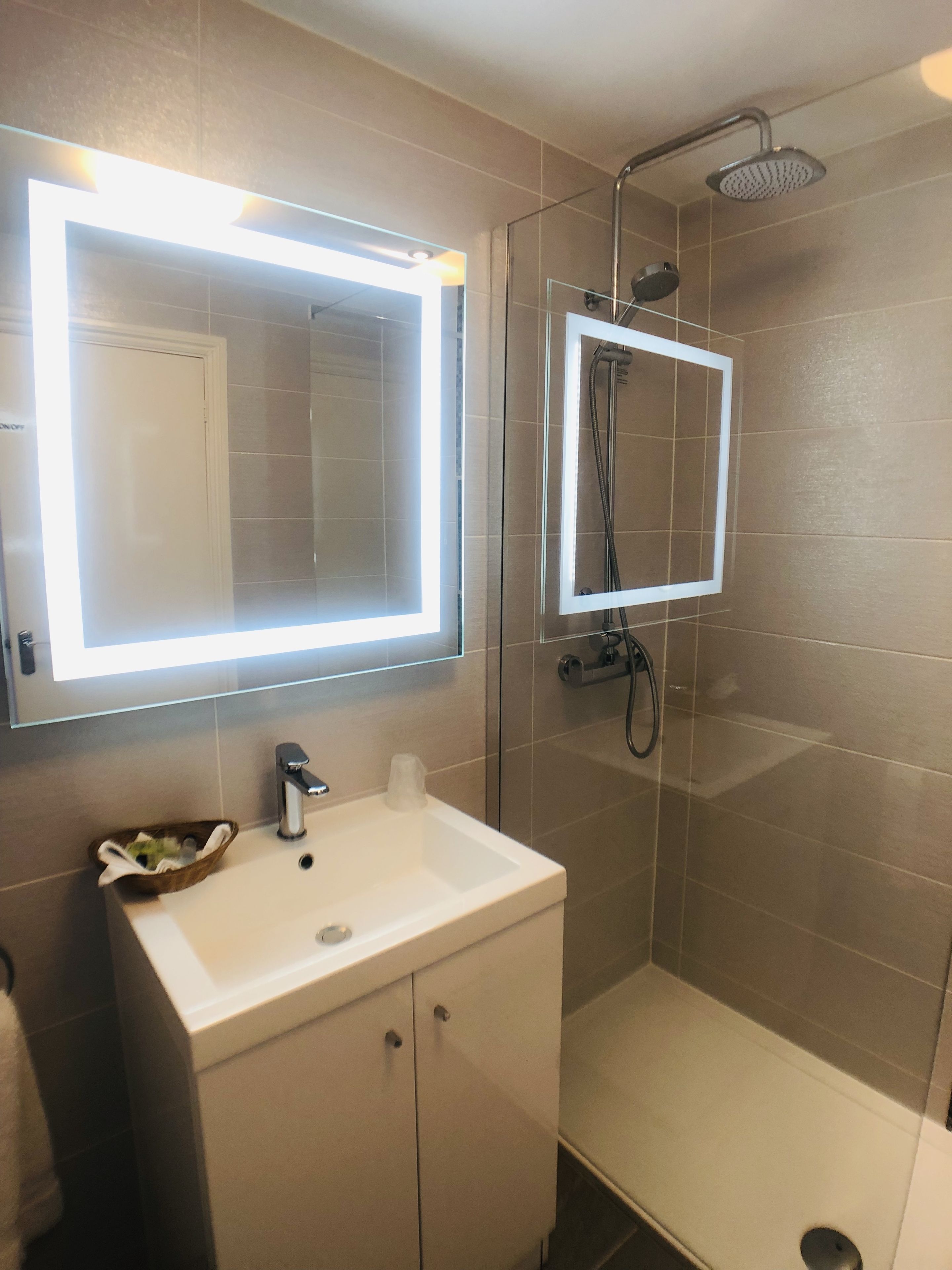 A bathroom with a white sink, a mirror with LED lights, a shower with a glass enclosure, and beige tiled walls.