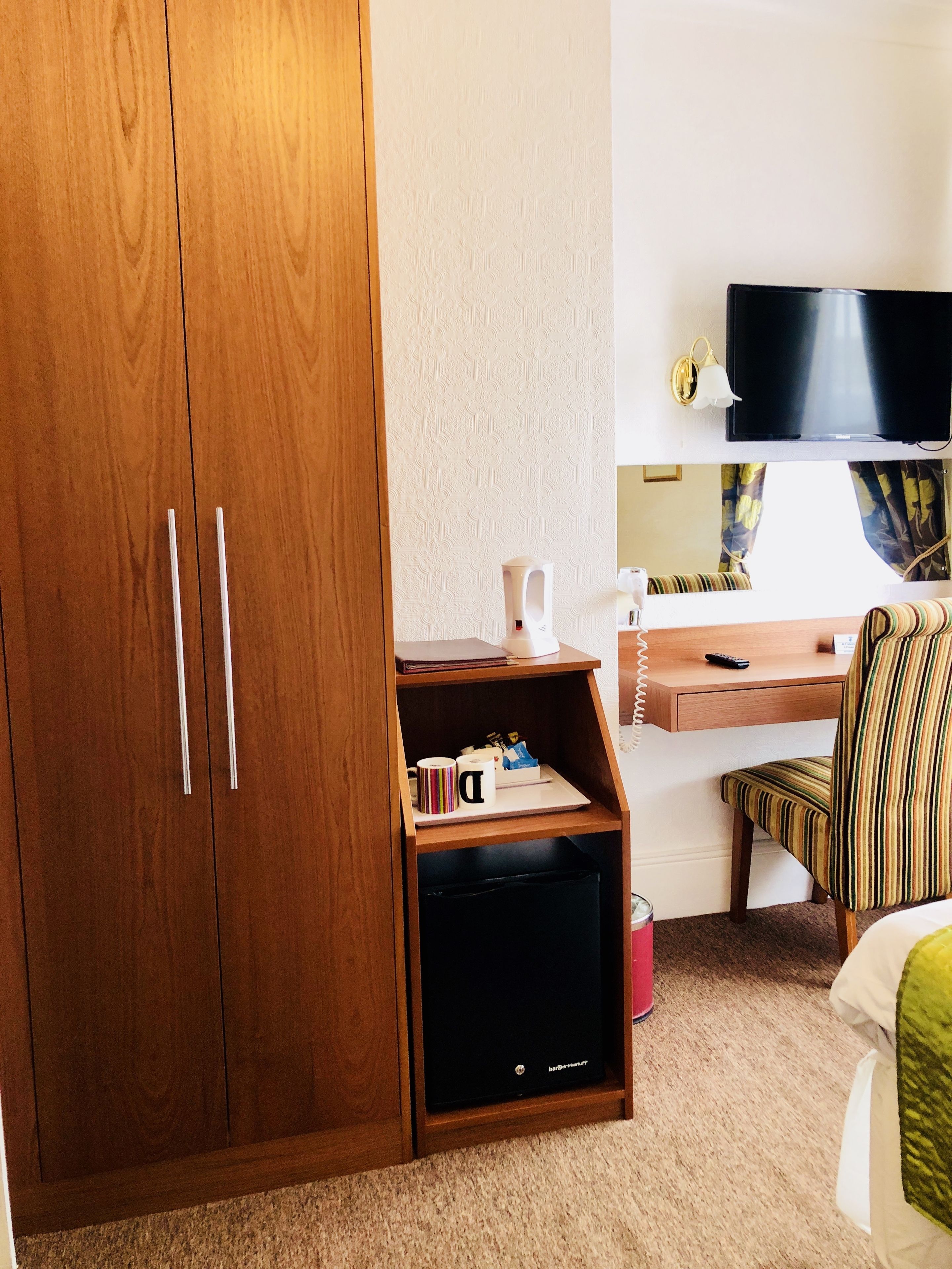 A hotel room with a wooden wardrobe, a desk with a chair, a television, and a small shelf with items.