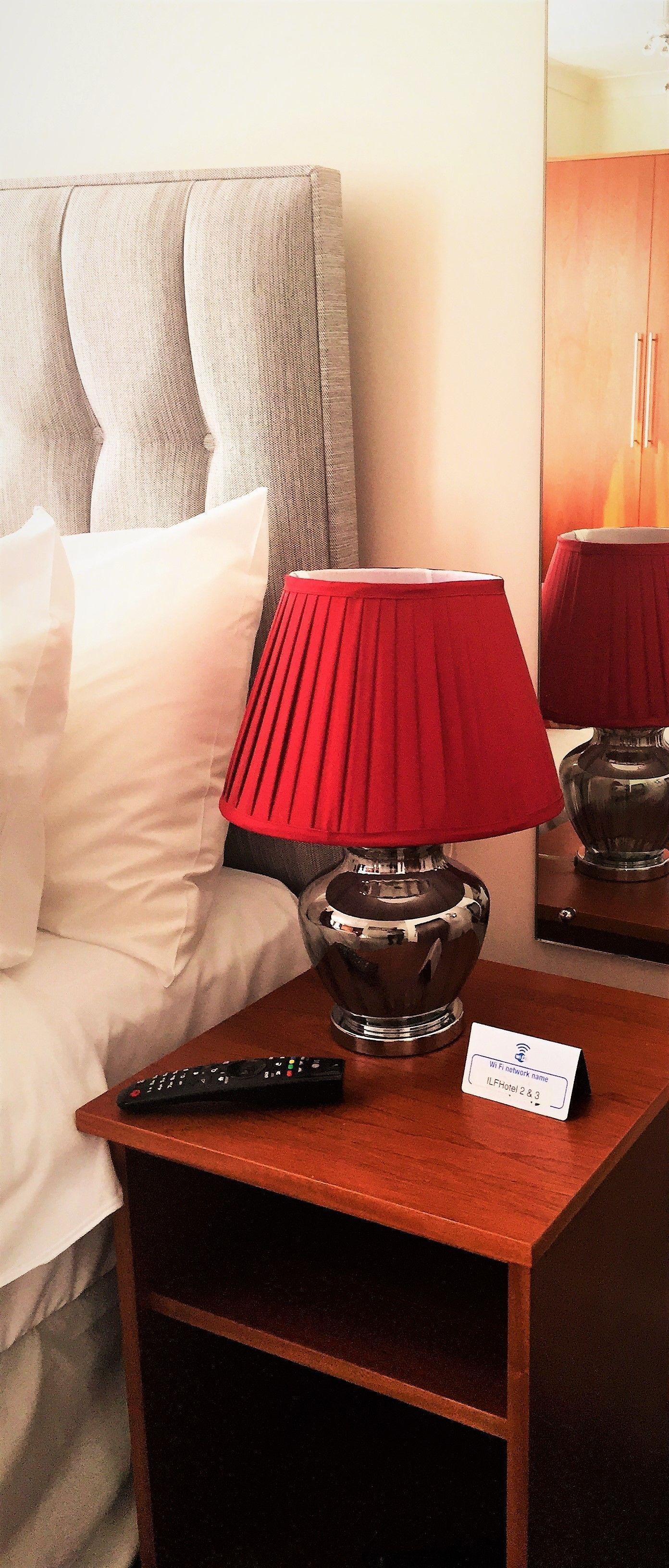 A hotel room with a bed, a red lamp, a remote control, and a Wi-Fi sign.