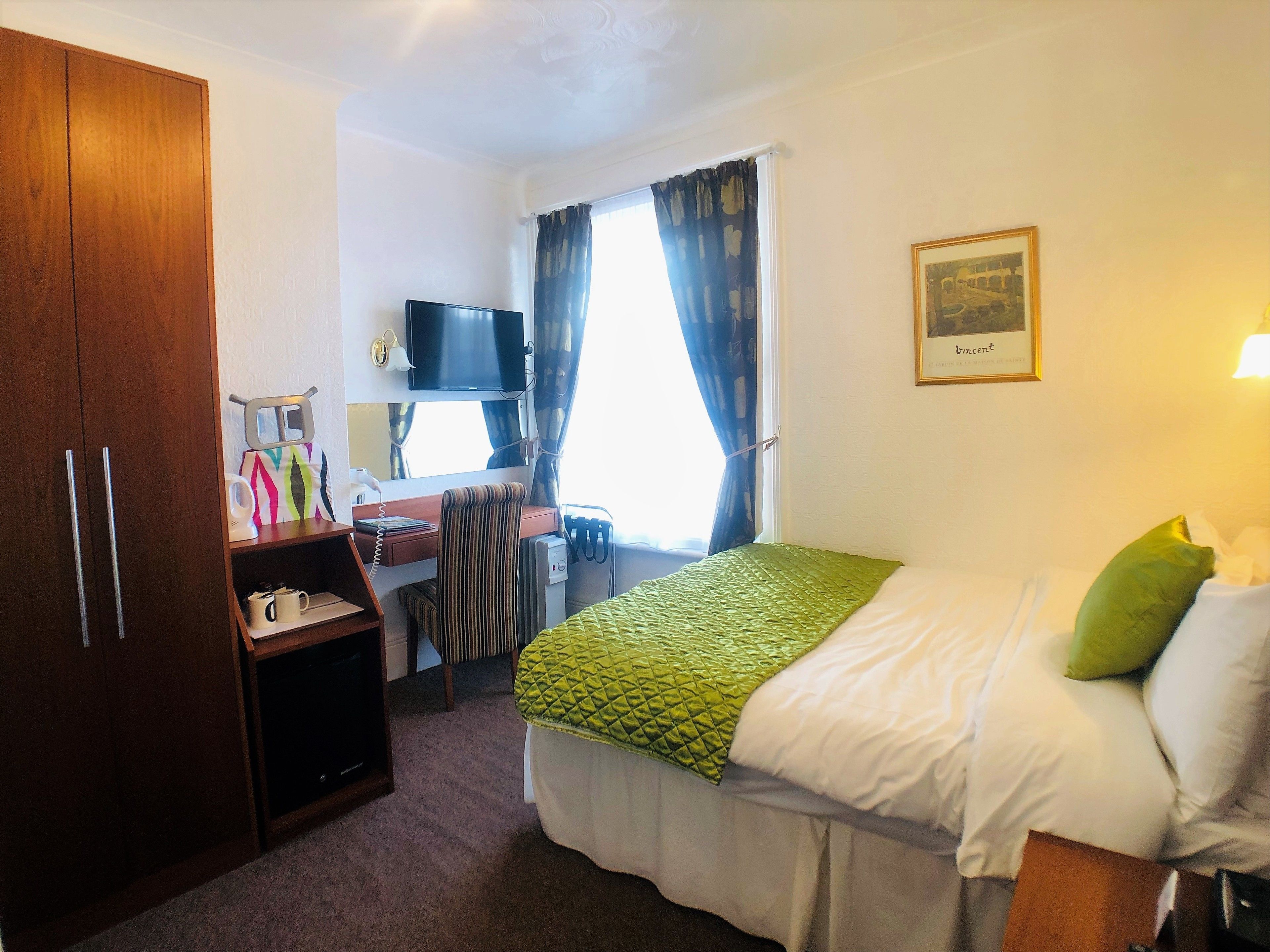 Habitación de hotel con cama, escritorio y silla, televisor, armario y ventana con cortinas.