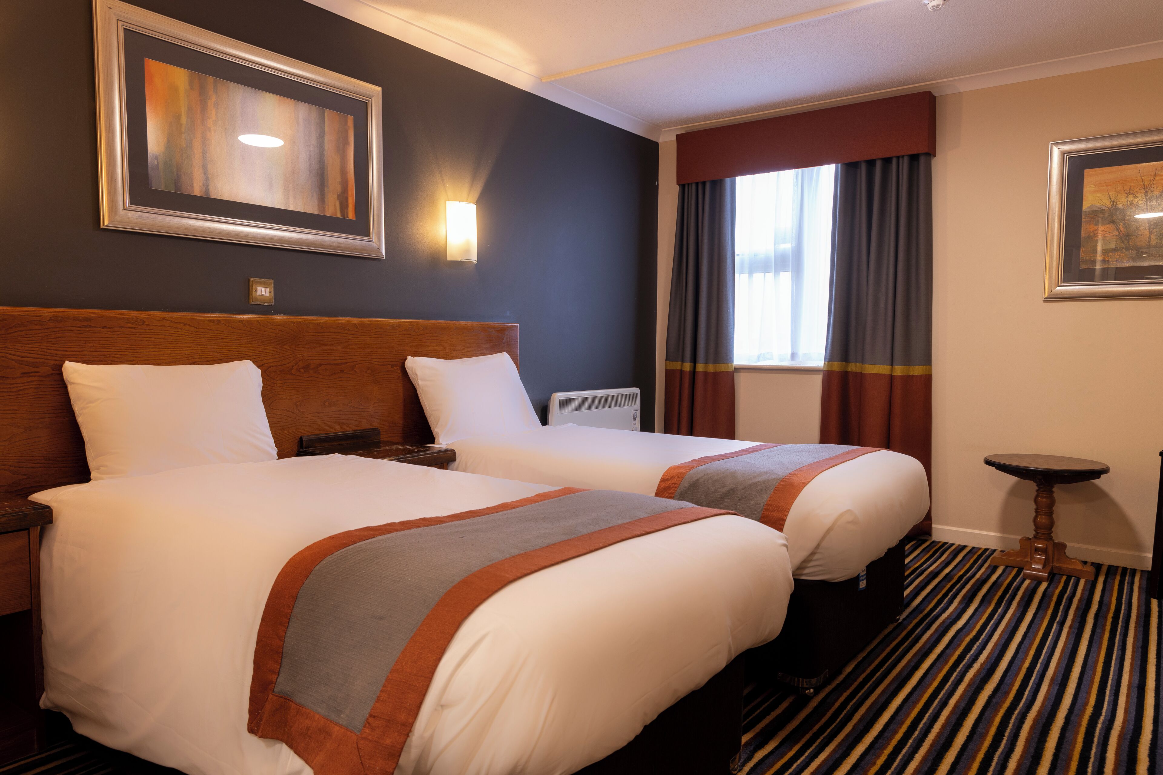 Ein Hotelzimmer mit zwei Betten, einem Nachttisch, einer Lampe, einem gerahmten Bild an der Wand und einem Fenster mit Vorhängen.