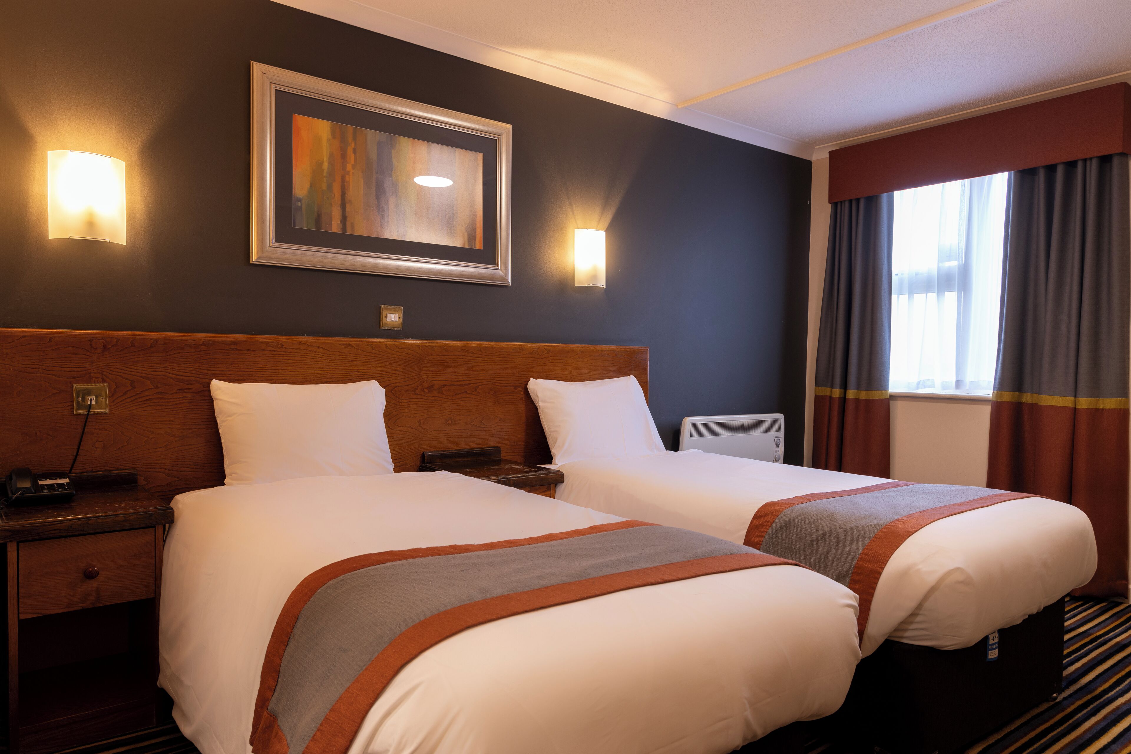 A hotel room with two beds, a wooden headboard, a framed artwork above the beds, a bedside table with a lamp, and a window with curtains.