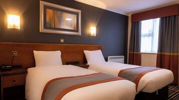 A hotel room with two beds, a wooden headboard, a framed artwork above the beds, a bedside table with a lamp, and a window with curtains.