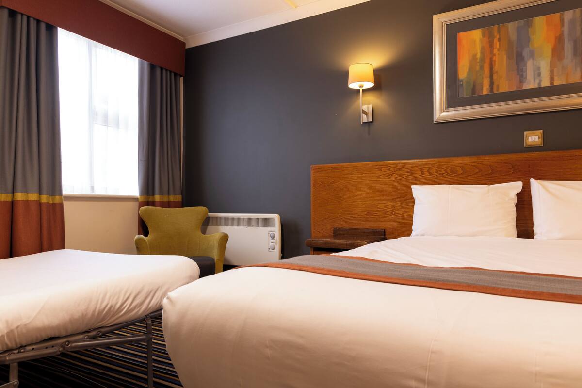 Ein Hotelzimmer mit zwei Betten, einem Holzkopfstück, einer Wandleuchte, einem Heizkörper und einem Fenster mit Vorhängen.