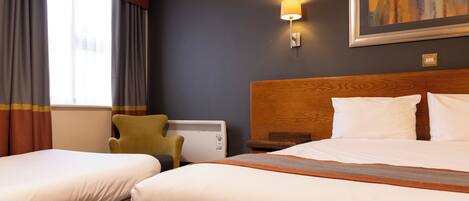 A hotel room with two beds, a wooden headboard, a wall-mounted lamp, a radiator, and a window with curtains.