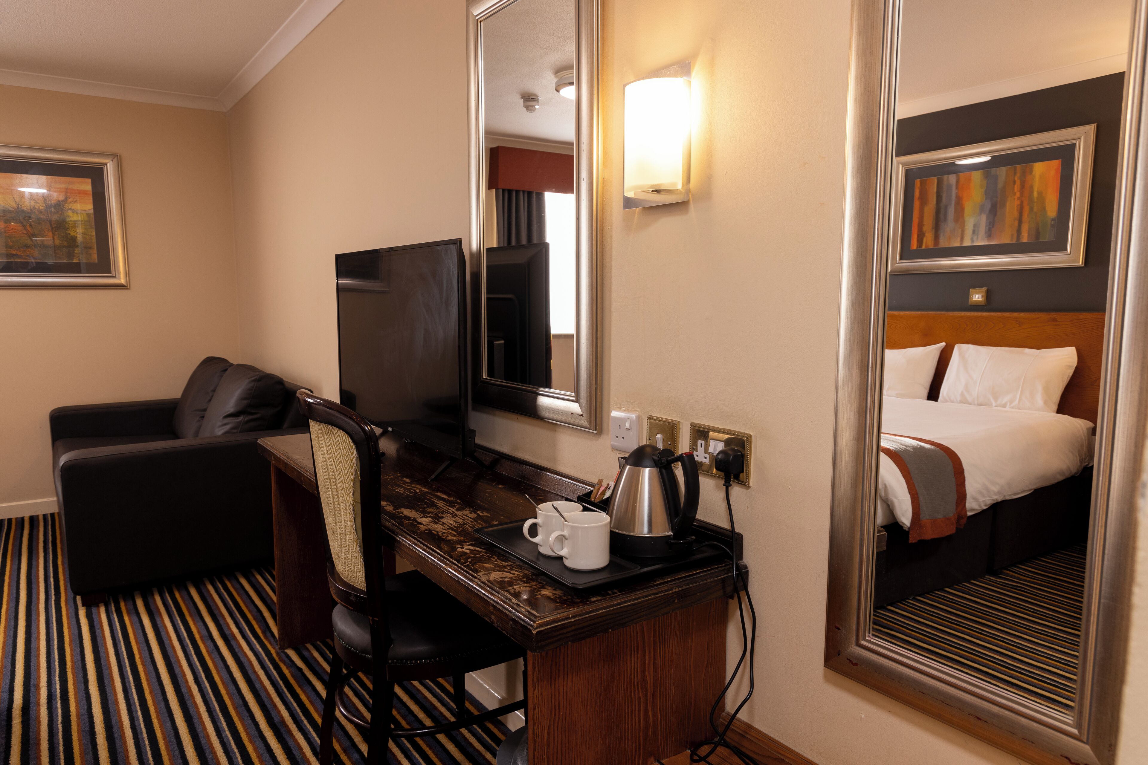 A hotel room with a bed, a desk with a coffee maker, a chair, a TV, and a striped carpet.