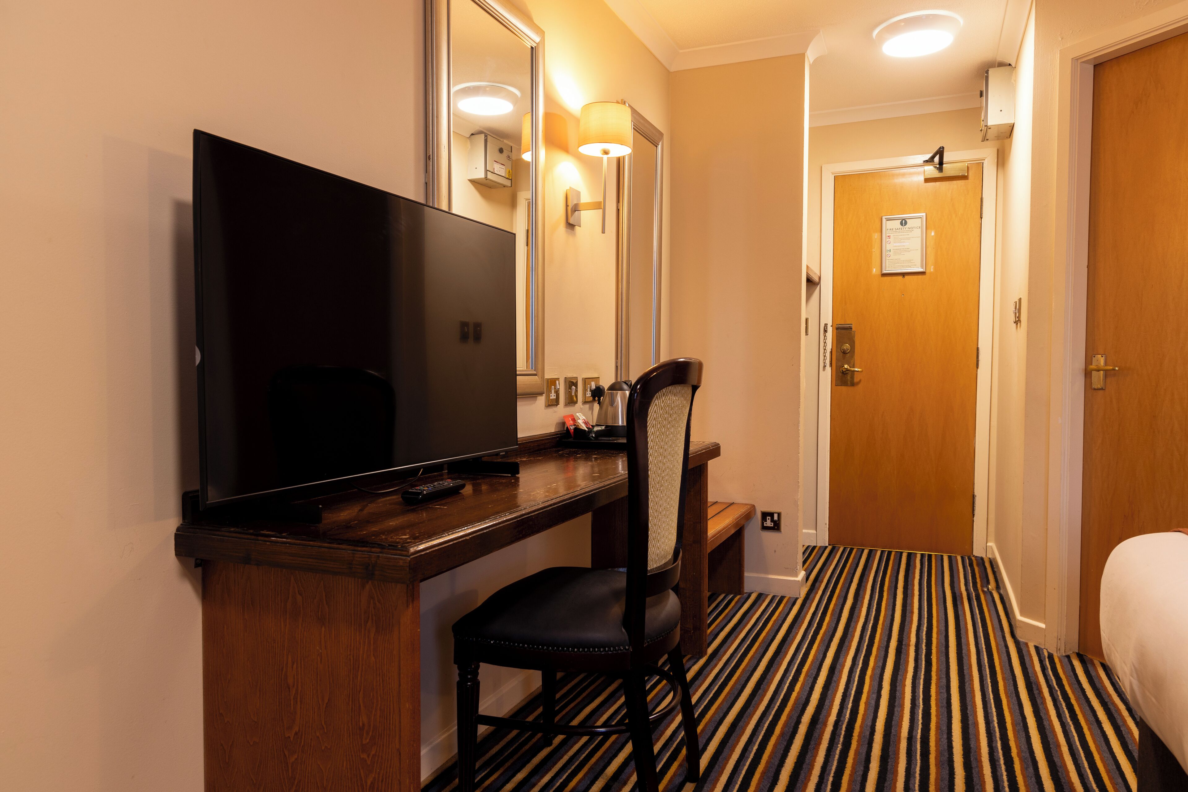 Une chambre d’hôtel avec un bureau en bois, une télévision, une chaise, un miroir, une porte et un tapis à rayures.