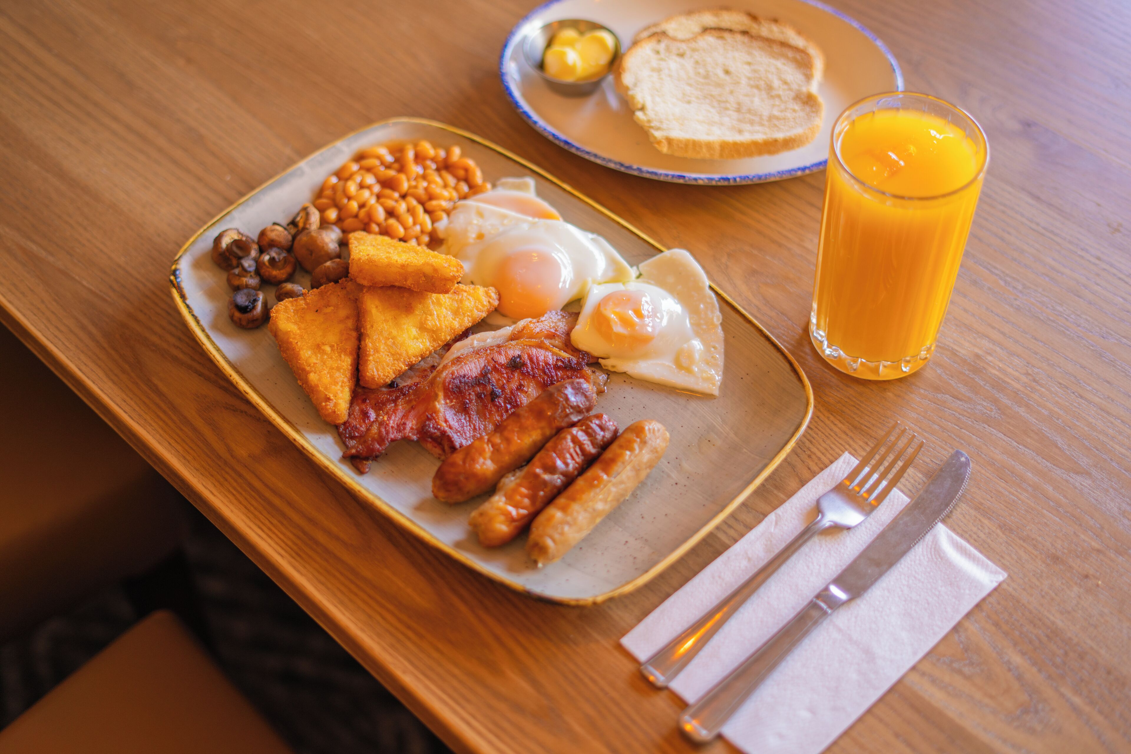 Un plato de desayuno con huevos, salchichas, frijoles, tostadas y un vaso de jugo de naranja sobre una mesa de madera.