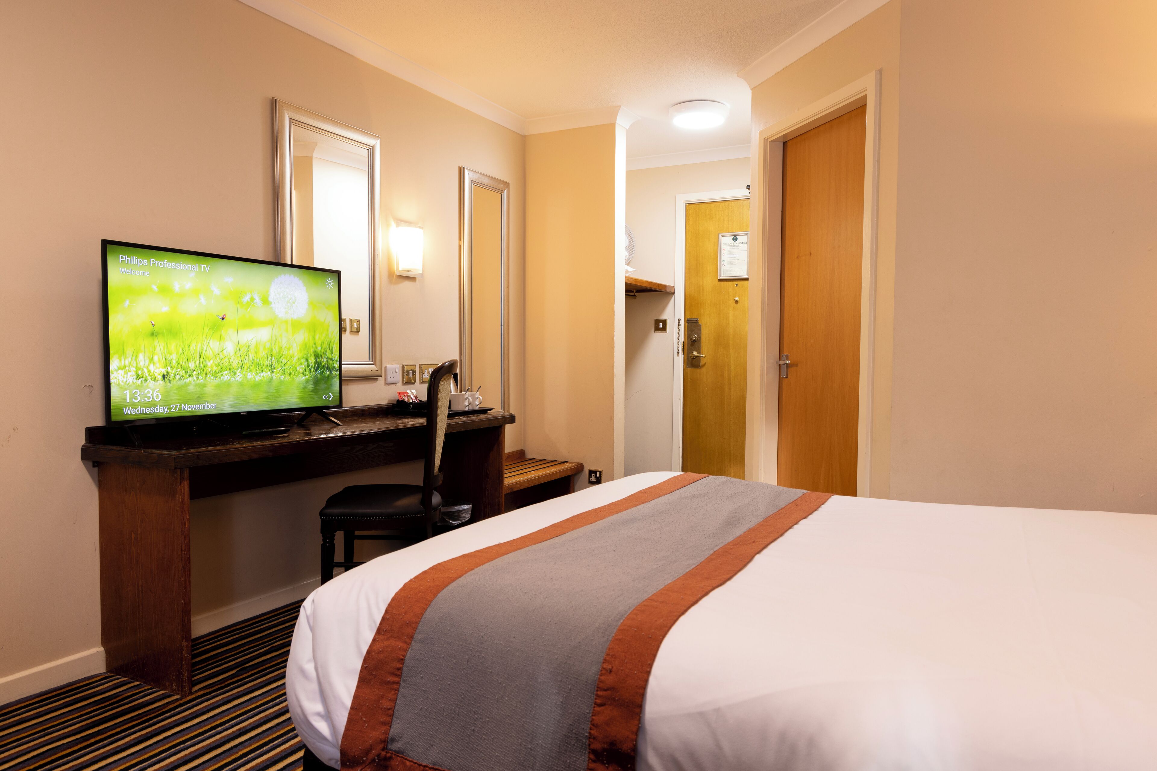 A hotel room with a bed, a desk with a television displaying a welcome screen, a chair, and a door leading to another room.