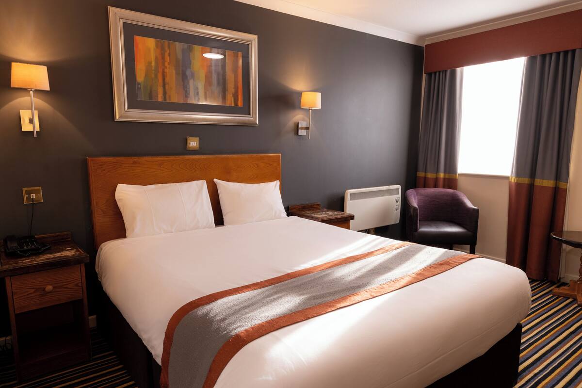 Ein Hotelzimmer mit einem großen Bett, zwei Nachttischen, einem Schreibtisch, einem Sessel und einem Fenster mit Vorhängen.