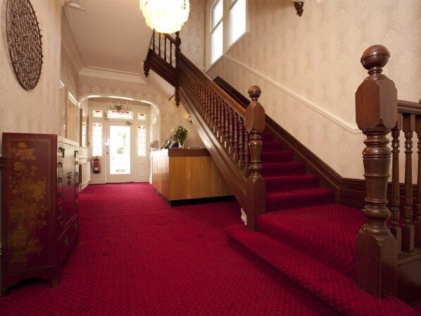 A grand staircase with red carpeted steps and wooden balusters, leading to a reception area with a wooden desk and a vase of flowers.