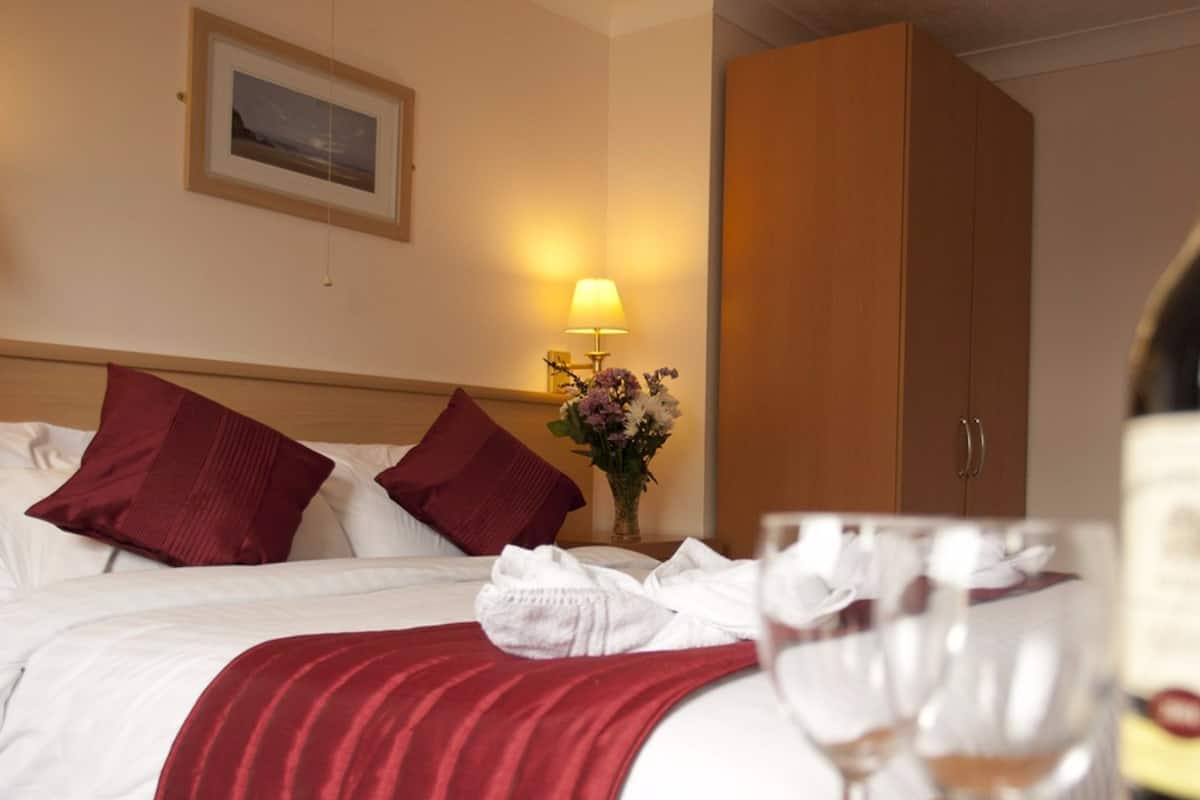 Ett hotellrum med en säng, kuddar, en sänglampa, en vas med blommor och ett vinglas.