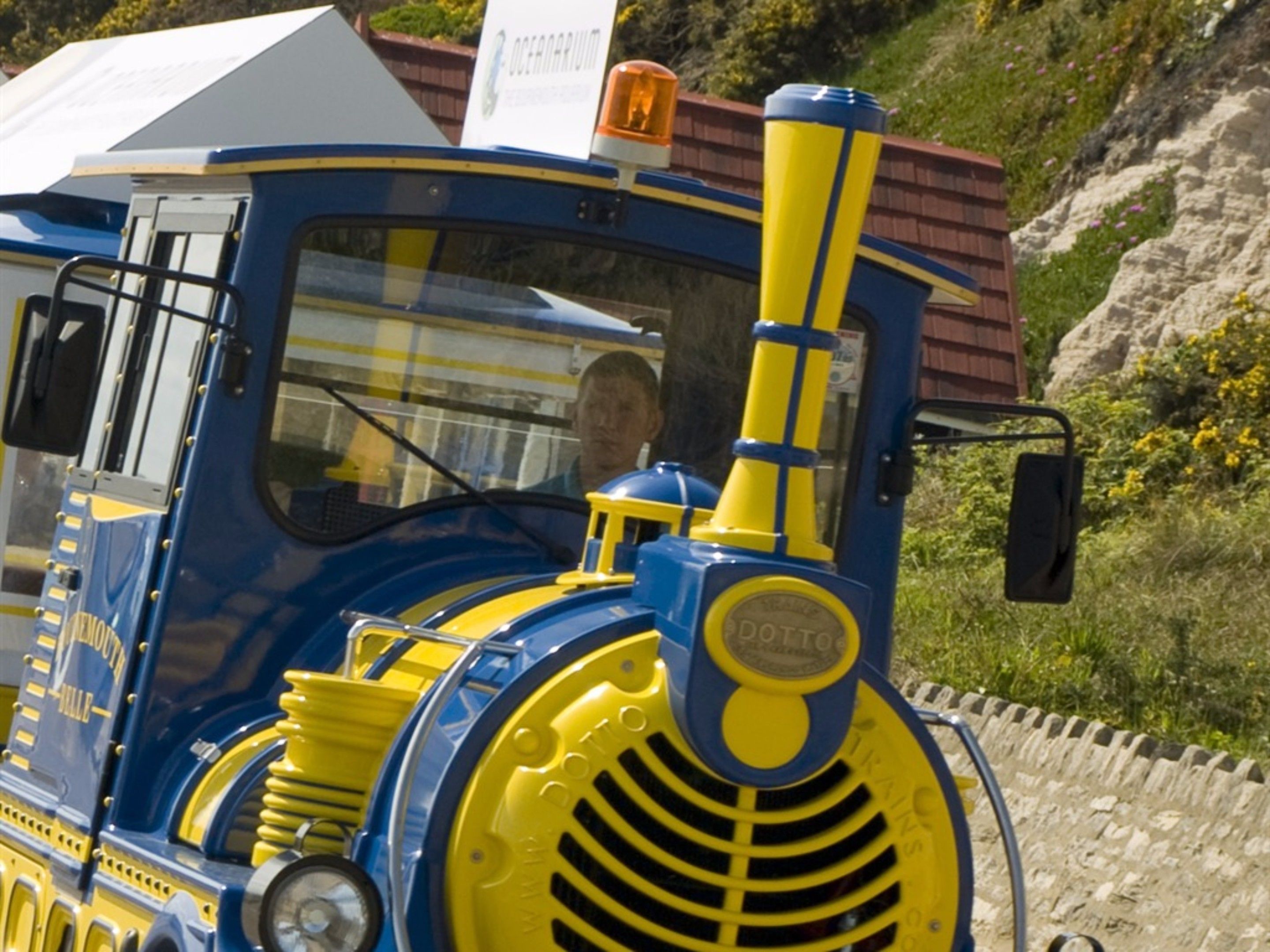 A vintage blue and yellow train engine with a driver’s cabin, set against a natural landscape with a stone wall and greenery.