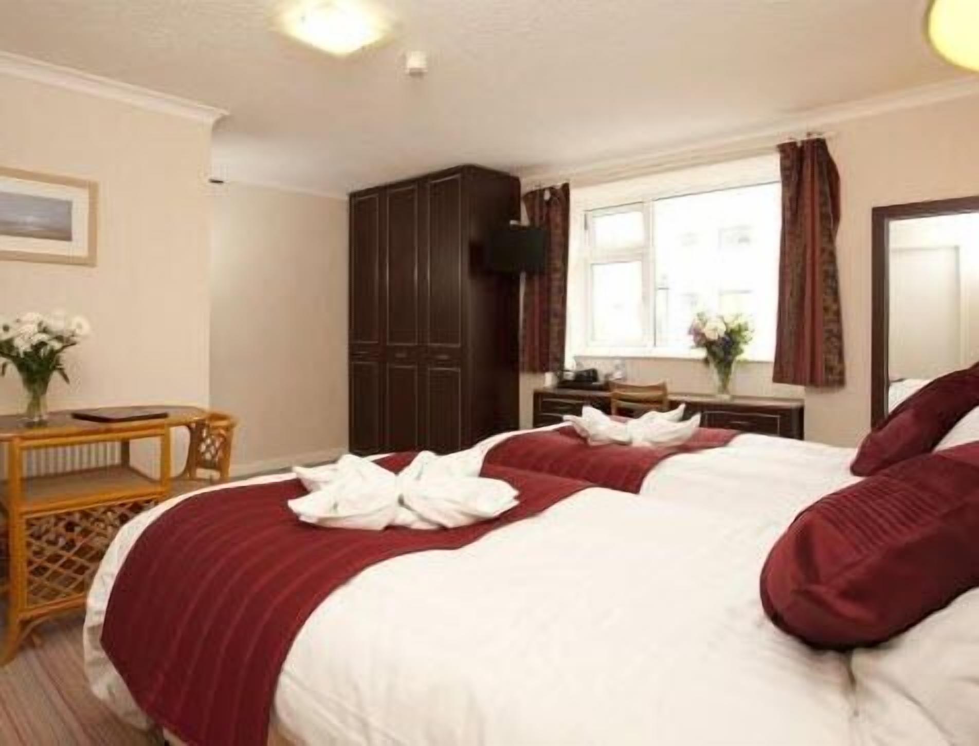 A hotel room with two beds, a wooden nightstand, a desk, a wardrobe, and a window with curtains.