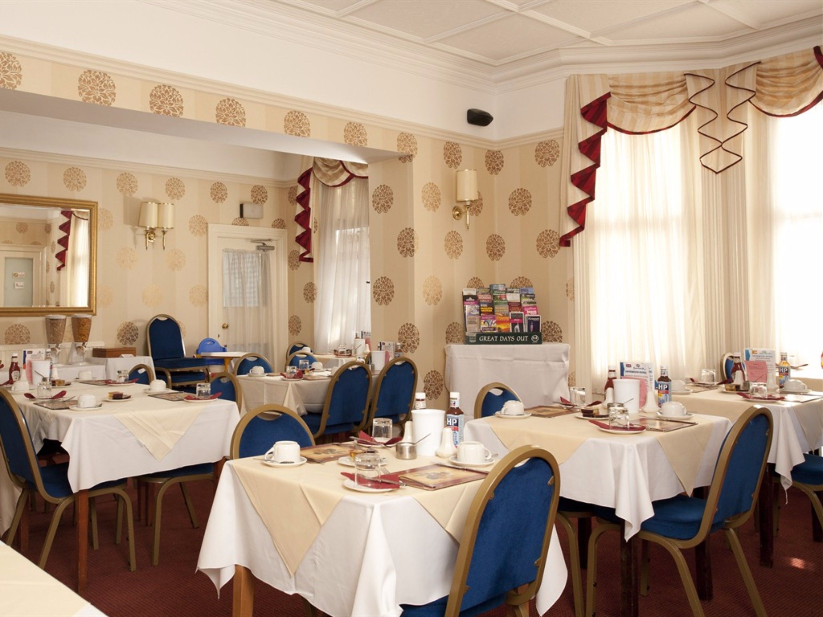 A formal dining room with multiple tables set for service, blue upholstered chairs, and a decorative wall with a mirror and patterned wallpaper.