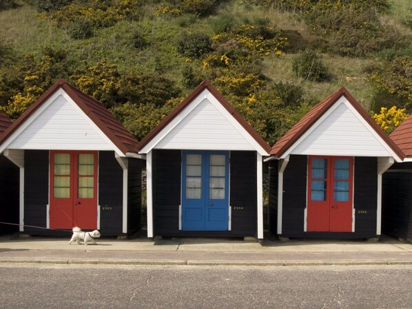 Una fila de tres cabañas de playa con puertas rojas y azules, numeradas 2363, 2384 y 2385, a lo largo de una carretera costera.