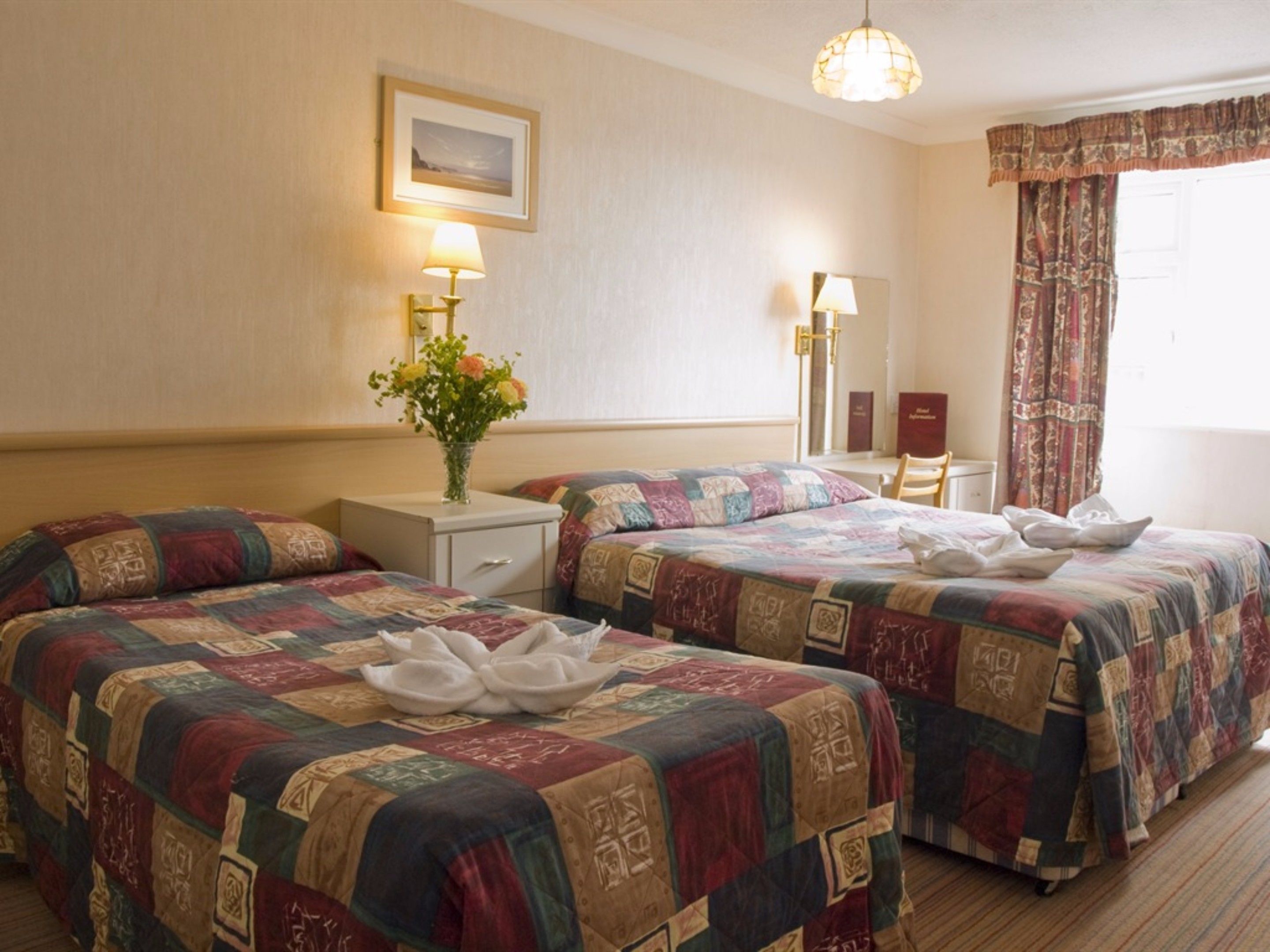A hotel room with two beds, a desk, a chair, a nightstand, a lamp, a vase with flowers, and a framed picture on the wall.