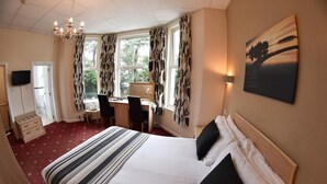 A hotel room with a large bed, a desk, and a chair. There is a television on the wall and a window with curtains. - The Mayfair Hotel (Bournemouth)