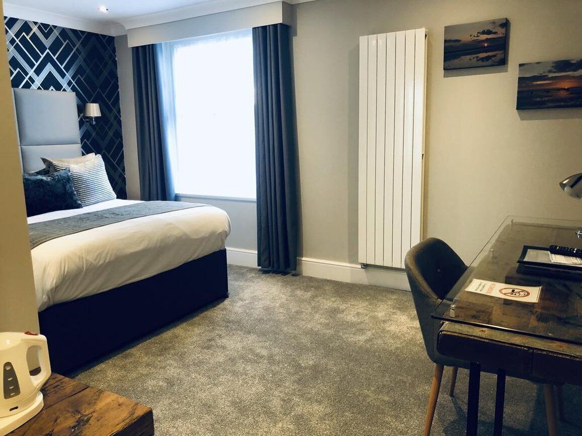 A hotel room with a bed, a desk, a chair, a radiator, and a window with curtains.