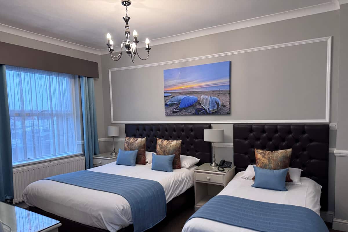 Habitación de hotel con dos camas, un cuadro grande de una playa, una araña y vistas al exterior a través de la ventana.