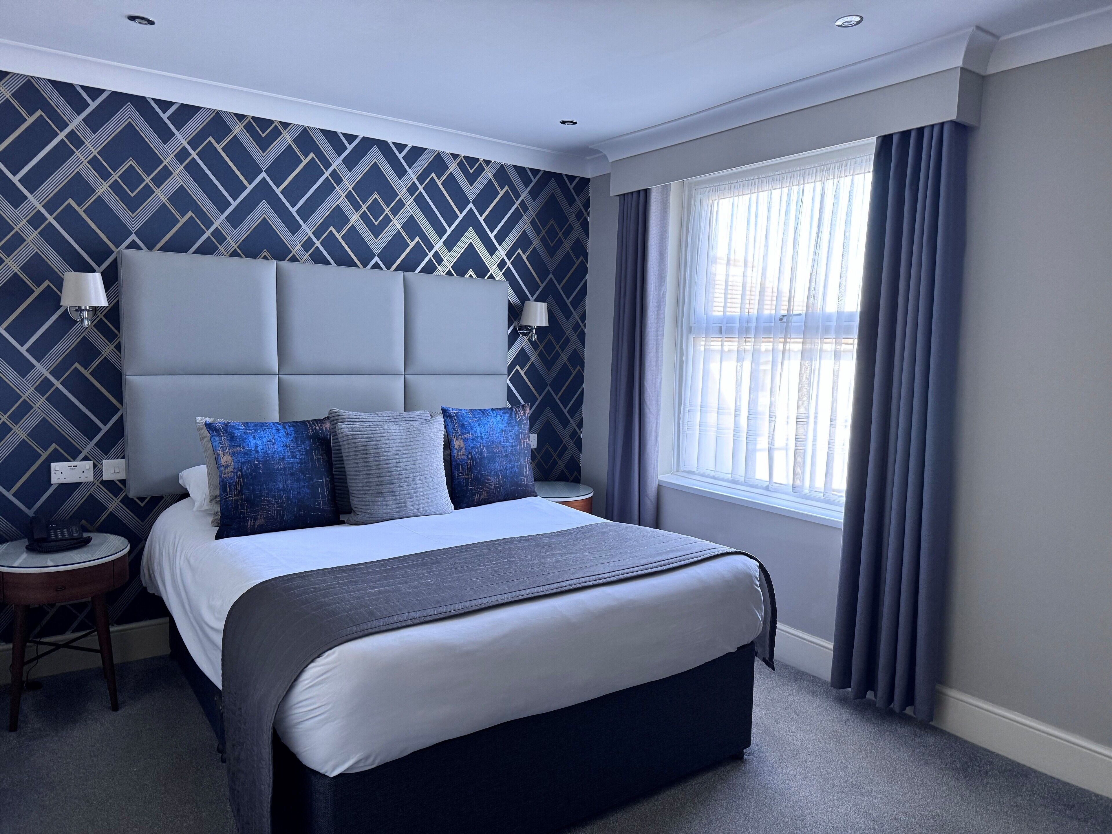 A modern hotel room with a large bed, a headboard, two bedside tables, and a window with curtains.