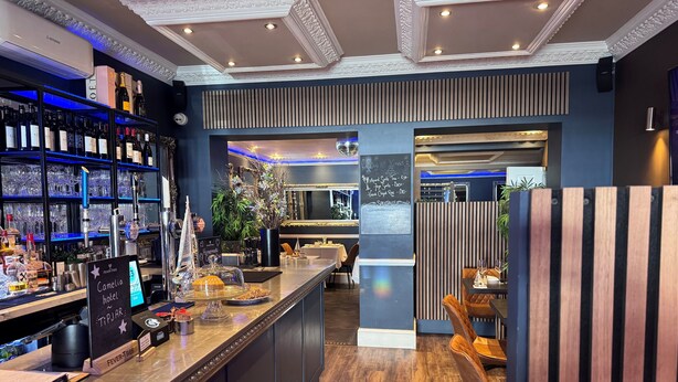 A bar area with shelves of bottles and glasses, a chalkboard menu, and a wooden counter. - Camelia Hotel (Southend-on-Sea)