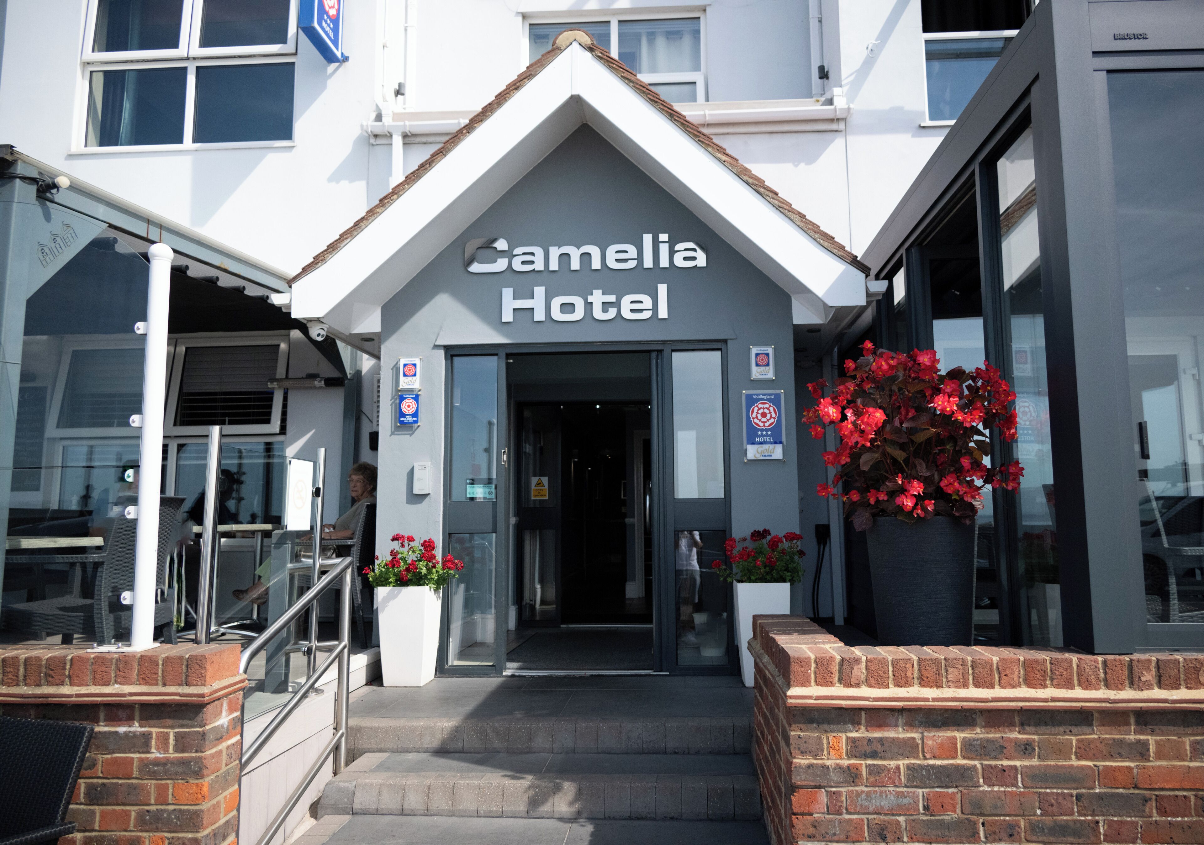 The entrance to the Camelia Hotel with a clear sign, steps leading up to the door, and a large potted plant with red flowers.