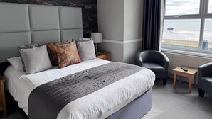 A modern hotel room with a large bed, a headboard with a marble-like design, a view of the sea through the window, and a small side table with a lamp. - Camelia Hotel (Southend-on-Sea)